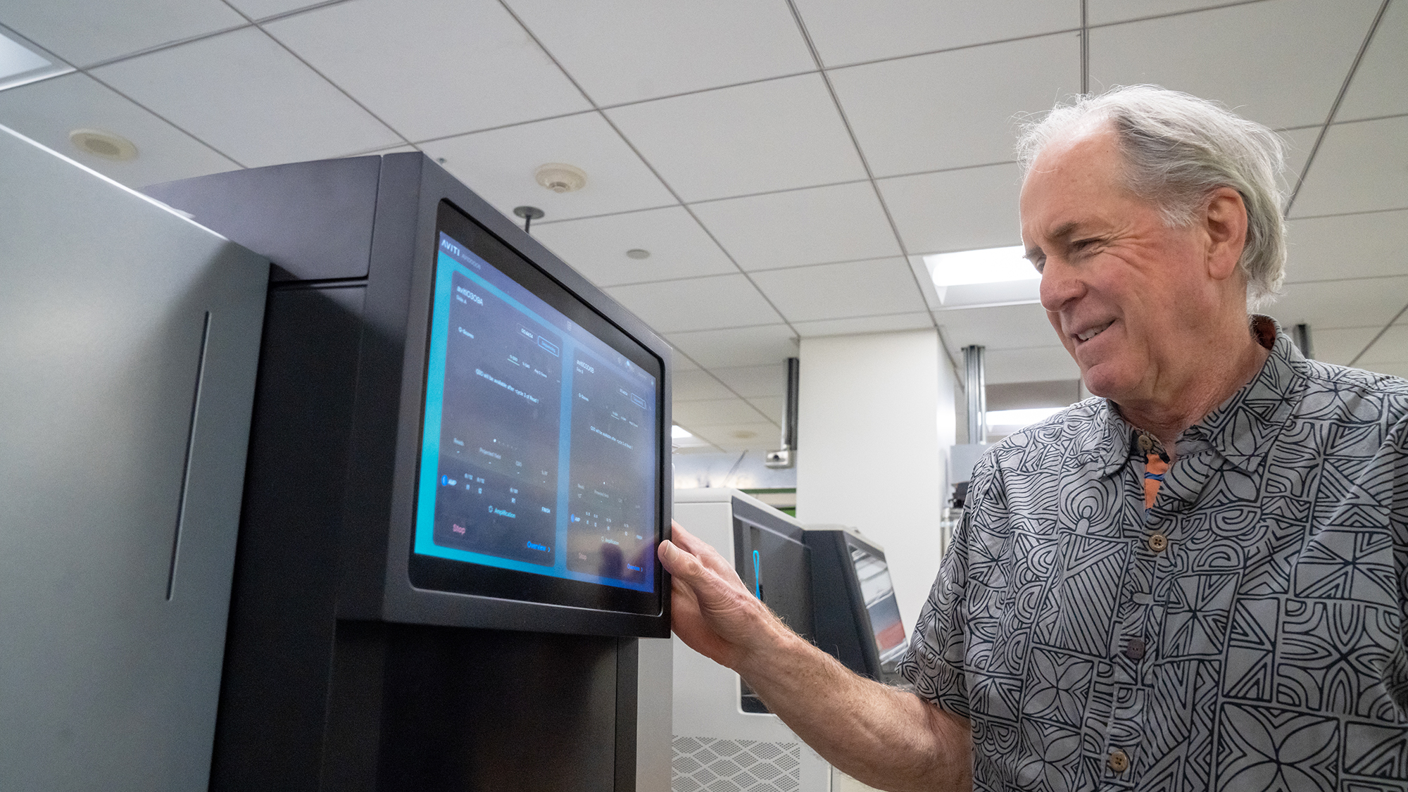 Richard Michelmore stands to the right of a black and blue screen on a genetic sequencing machine, looking at the screen.