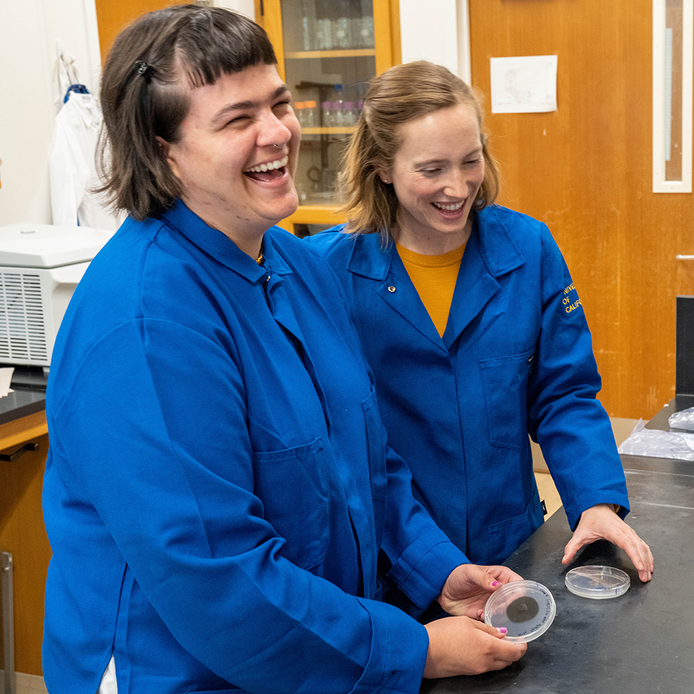 Two female researchers in blue lab coats laughing. 