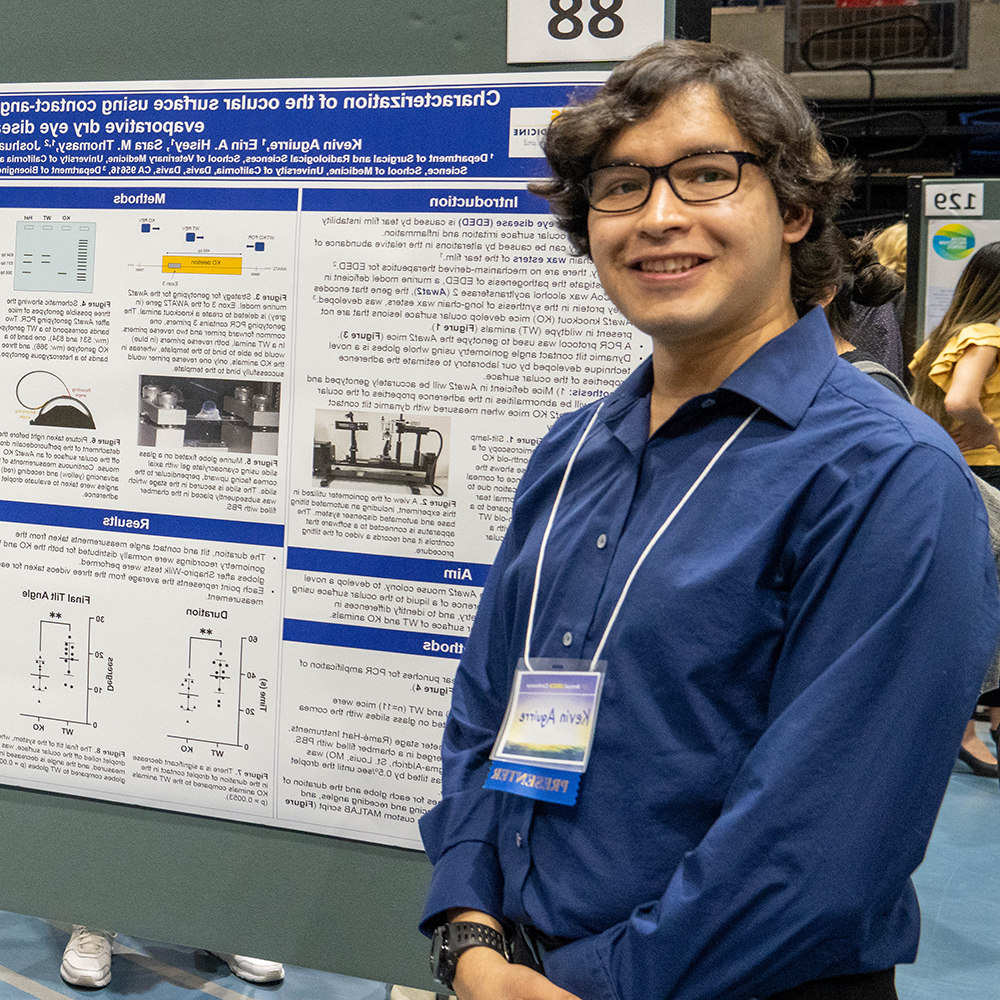Male student in blue shirt standing in front of his research poster smiling.