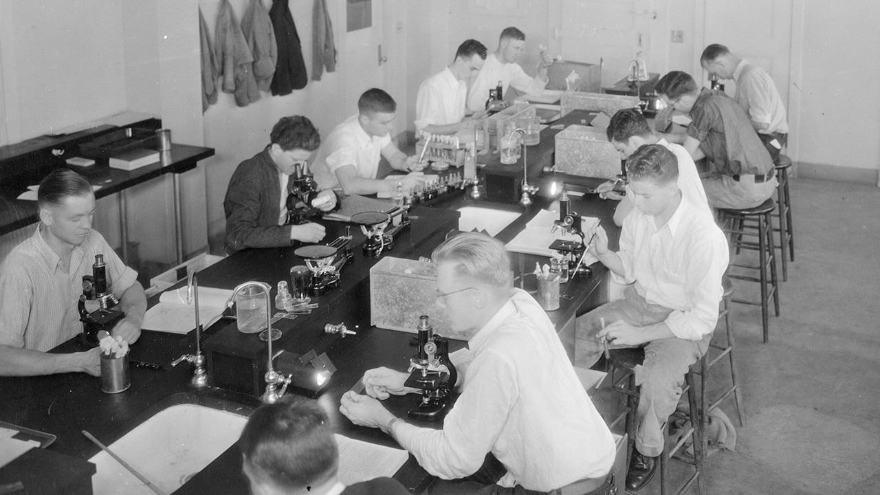 A black and white archival photograph of male students seated along a long laboratory table with microscopes. 