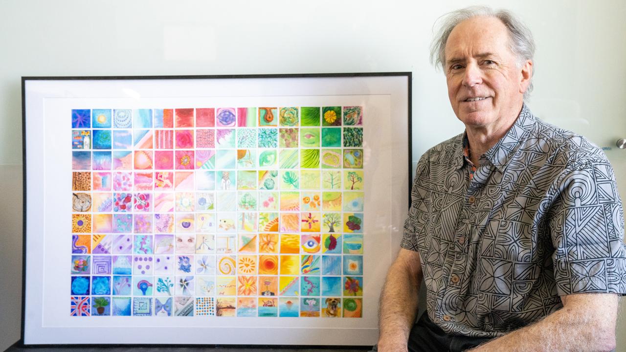 Richard Michelmore is seated to the left of a framed piece of art made of a grid of multicolored squares.