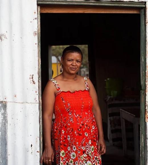 A woman with dark skin standing in the doorway of her home wearing a red garment.