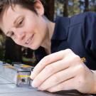 A person with short hair wearing a dark polo shirt and a green watch is engaged in an outdoor activity, working with a small, clear plastic device that appears to be a scientific instrument on top of a metallic surface. The setting includes trees and natural light, suggesting the activity is taking place in a wooded area during the day.