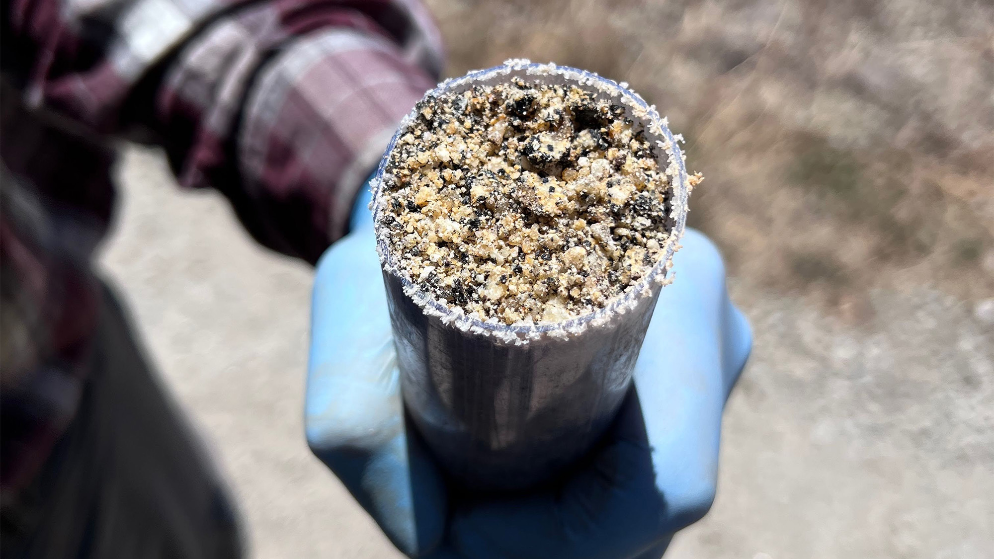 Blue-gloved hand holding clear tube filled with sandy soil and gravel core