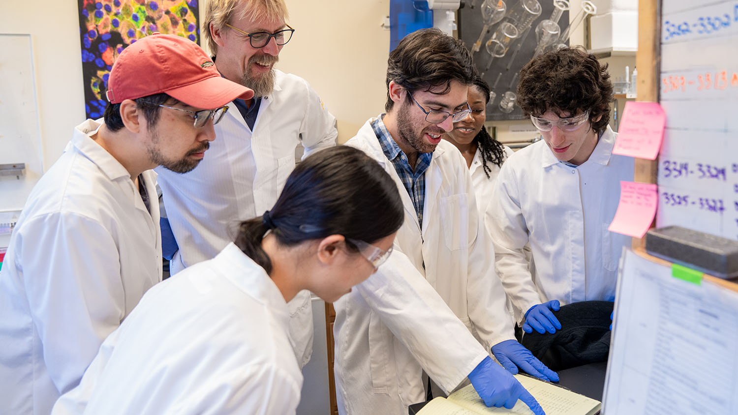 Group of researchers in lab coats gather around a bench reviewing notes with one person pointing at a notebook