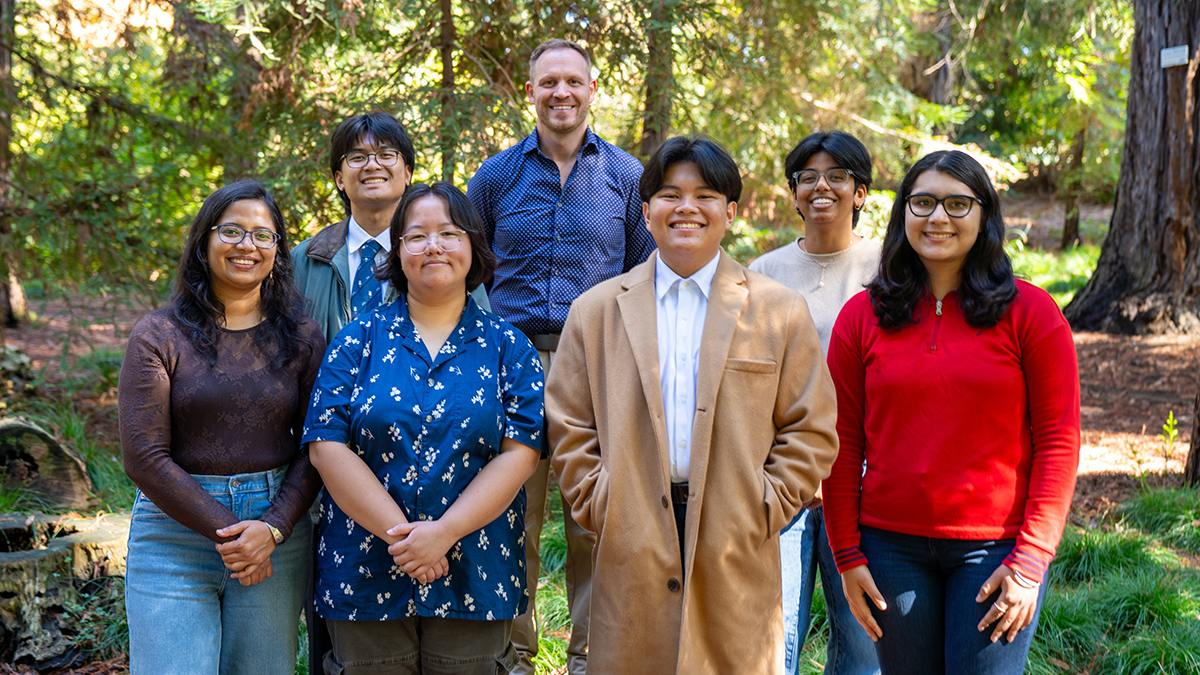 Group of students and instructor stand smiling together in the arboretum surrounded by trees and greenery.