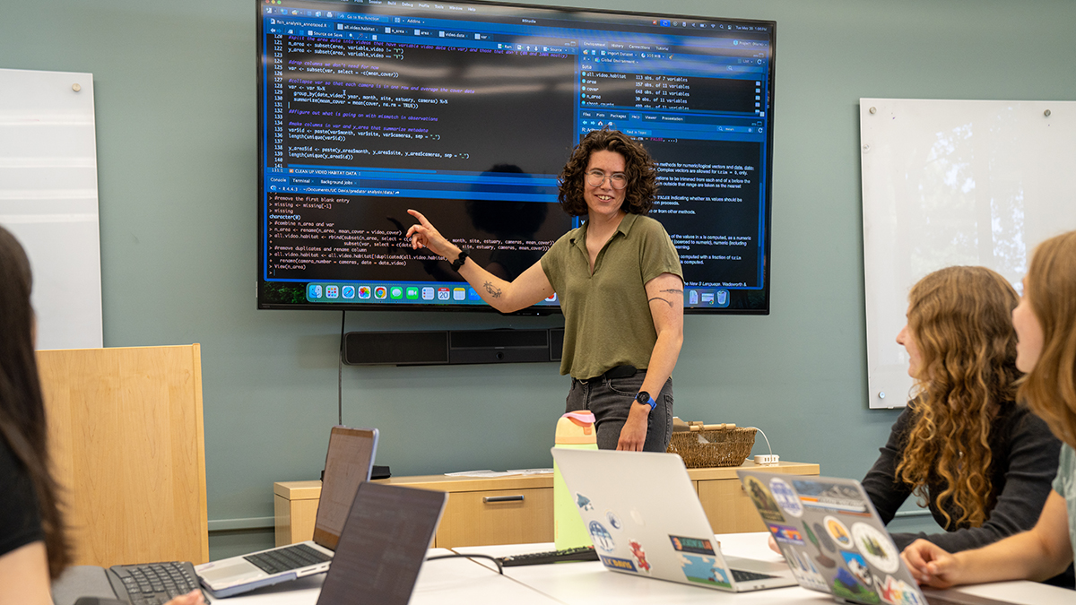 Claire Murphy stands at the front of a classroom pointing at a large screen displaying code, while undergraduate students seated at a table with laptops look on.