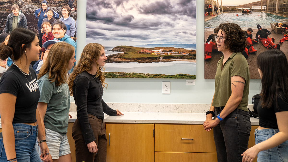 Claire Murphy (right), wearing a green shirt, stands and speaks with five undergraduate students in a room at the Bodega Marine Laboratory. Behind them are large photos of marine life, a coastal research facility, and students in wetsuits, emphasizing the lab’s immersive, hands-on learning environment.