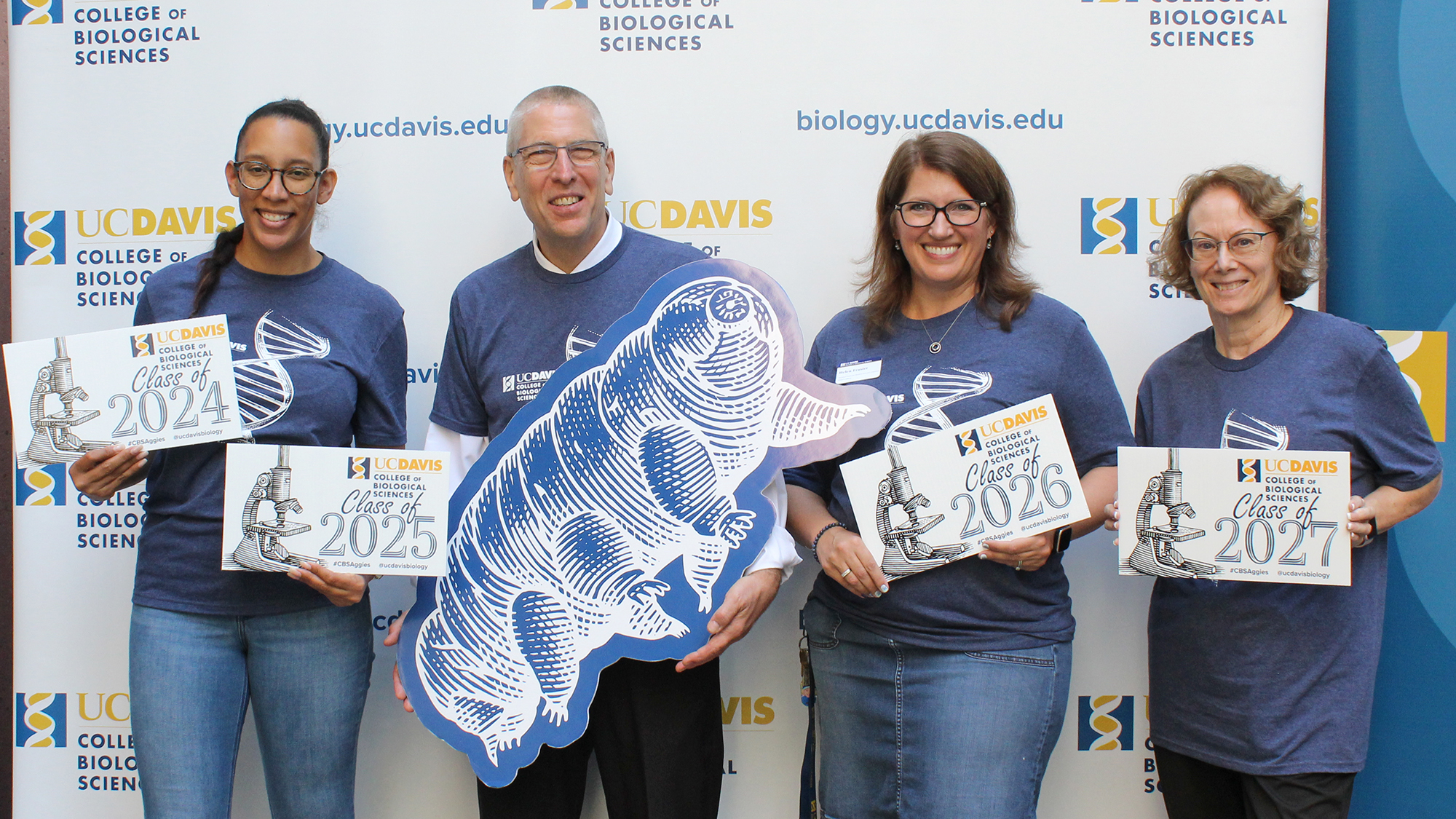 Four UC Davis College of Biological Sciences staff and faculty smile in front of a branded backdrop while holding class year signs (2024–2027) and a large cutout of a blue woodcut-style tardigrade. All are wearing matching navy blue CBS shirts featuring a DNA helix design.