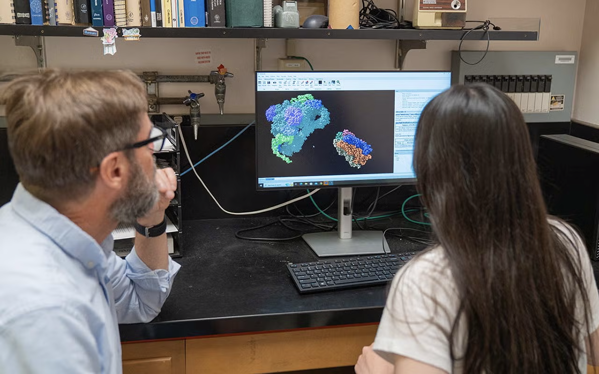 Two people sit at a lab bench, closely examining a colorful 3D molecular model displayed on a computer screen. The lab setting includes books, lab equipment, and coiled cables, indicating a research environment focused on molecular or structural biology.