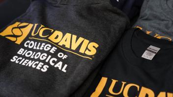 A variety of UC Davis College of Biological Sciences apparel laid out on a table, featuring dark-colored shirts and sweatshirts with the college's logo and name printed in white and gold.