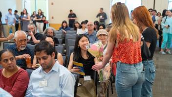 Students and families at an awards ceremony. One student receives flowers and smiles, surrounded by seated attendees in a packed room.