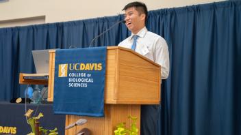 Student speaker at podium with UC Davis College of Biological Sciences banner, smiling and addressing an audience with enthusiasm.