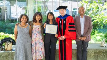 Award recipient poses with certificate and family. Group stands outside in sunlight with greenery and academic building in background.