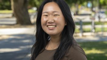 Person smiling outdoors on a tree-lined campus walkway, wearing a brown short-sleeve top with long dark hair.