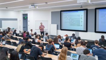 University lecture hall with instructor presenting slides to seated students.