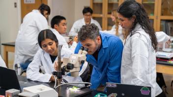 Diverse students in lab coats gathered around a microscope in a classroom lab