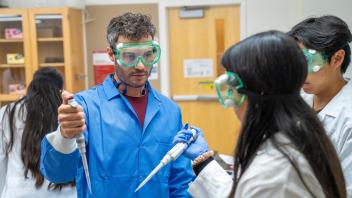 Teacher in green goggles and blue gloves demonstrating a pipette to students in a lab