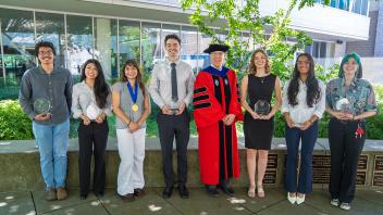 Eight people smiling on campus lawn, center in red academic gown holding awards and flowers
