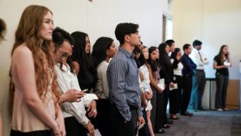 Students line the wall, standing respectfully with programs in hand. Ceremony in progress, many appearing attentive and professionally dressed. 