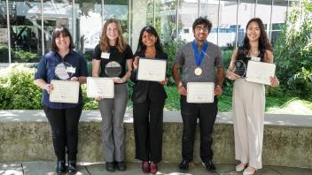 Five students stand proudly outdoors holding certificates and awards, smiling for the camera in front of a glass building.