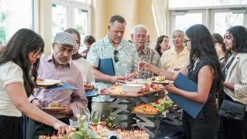 Guests line up at a reception buffet, filling plates with sandwiches, vegetables, and snacks in a bright indoor space.