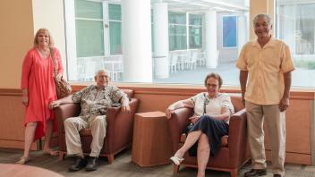 Four smiling guests, seated and standing, relax in a sunny lobby area with wide windows and casual conversation.
