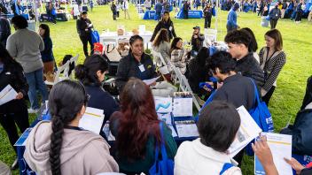  A large group of attendees gathers at a booth under a canopy tent, speaking with representatives and examining brochures. The surrounding grassy field is filled with similar booths and attendees walking around.