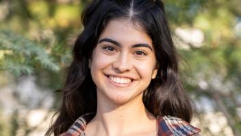 A person with long dark hair smiling and standing in front of a tree