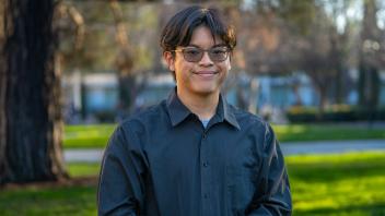 A person in a light blue shirt and glasses standing outside in front of a row of trees
