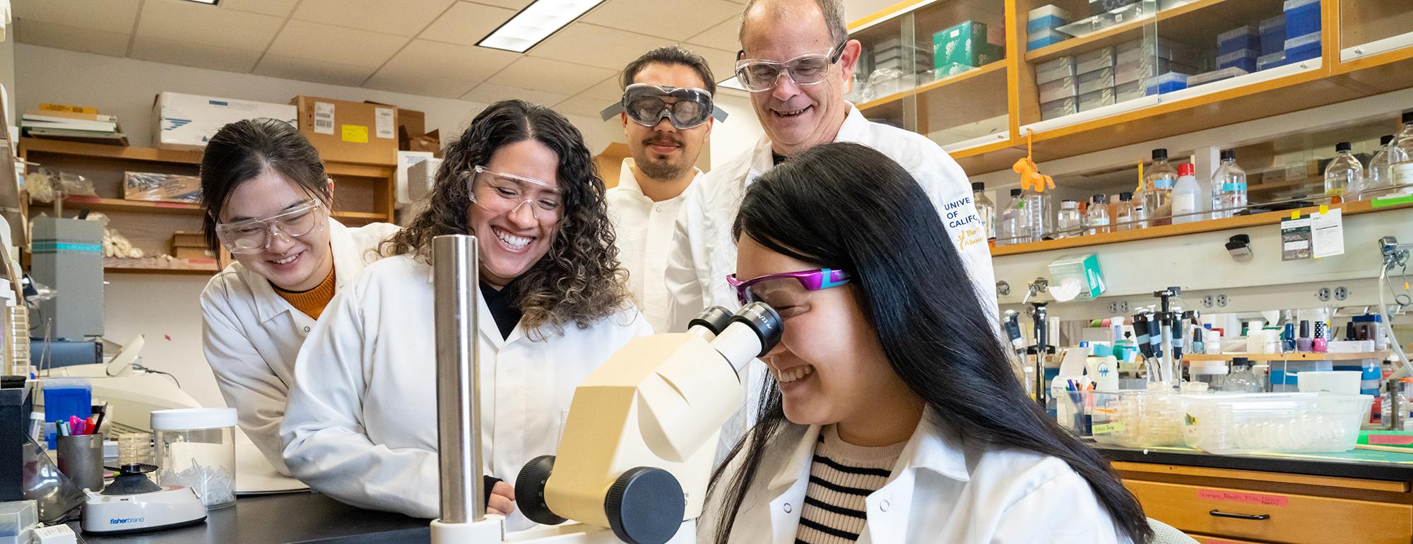 Five researchers in lab coats and safety goggles gather around a microscope in a laboratory. One person is looking into the microscope while others observe and smile, surrounded by shelves and lab equipment.
