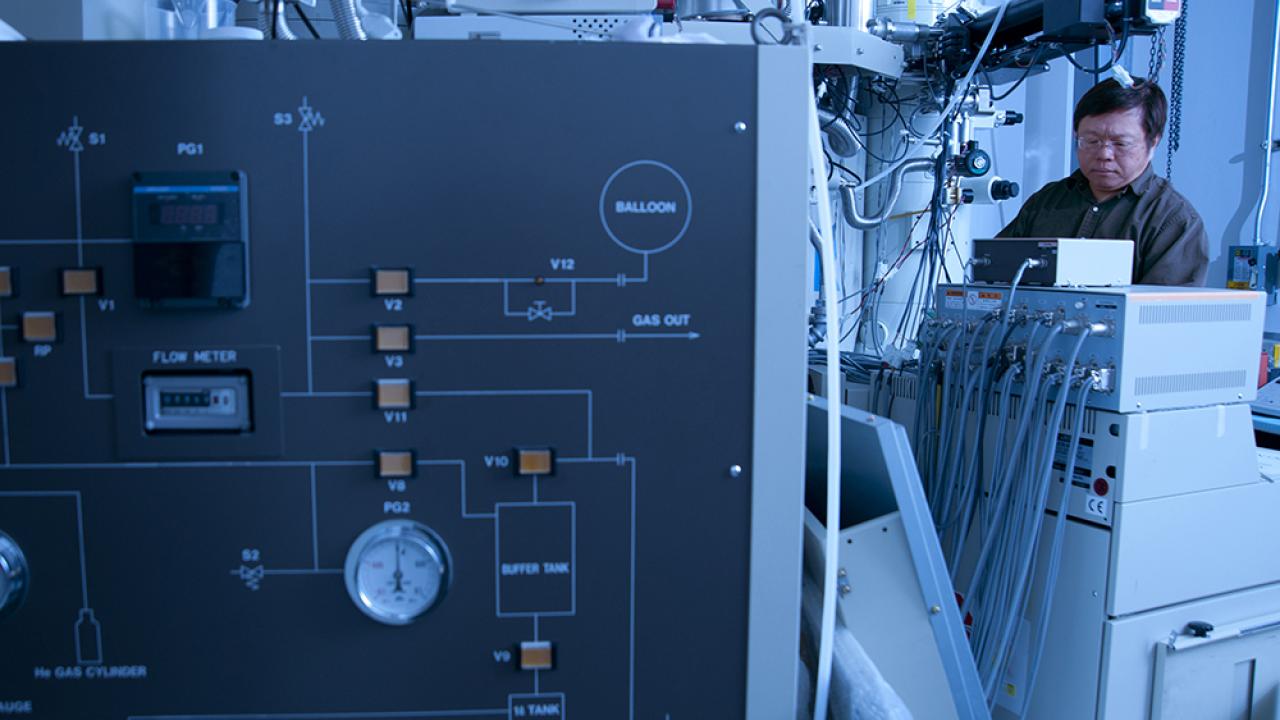 A control panel featuring labels such as ‘BALLOON,’ ‘GAS OUT,’ and ‘FLOW METER,’ along with various gauges, indicators, and switches. In the background, a person is seated in front of multiple computer monitors. The setting appears to be a technical or scientific environment.