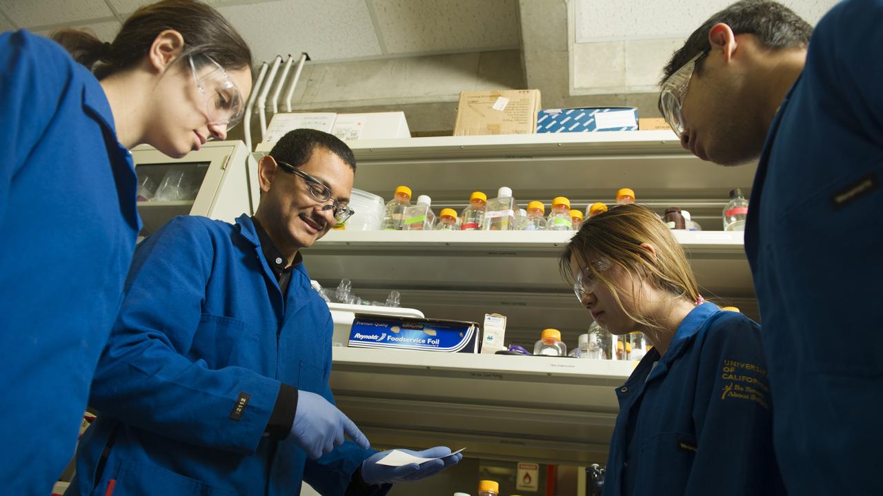 A scientist in a blue lab coat and gloves explains a procedure to three students in a research lab. All are wearing safety goggles and blue UC-branded lab coats, standing in front of shelves lined with bottles and lab supplies.
