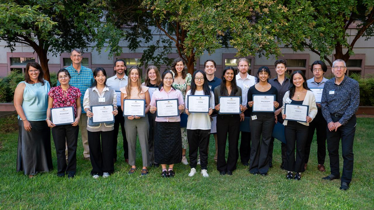 A group of sixteen individuals standing outdoors, each holding a certificate, with trees and a building in the background. They are dressed in business-casual attire, smiling for the photo, and are arranged in a single row on the grass.