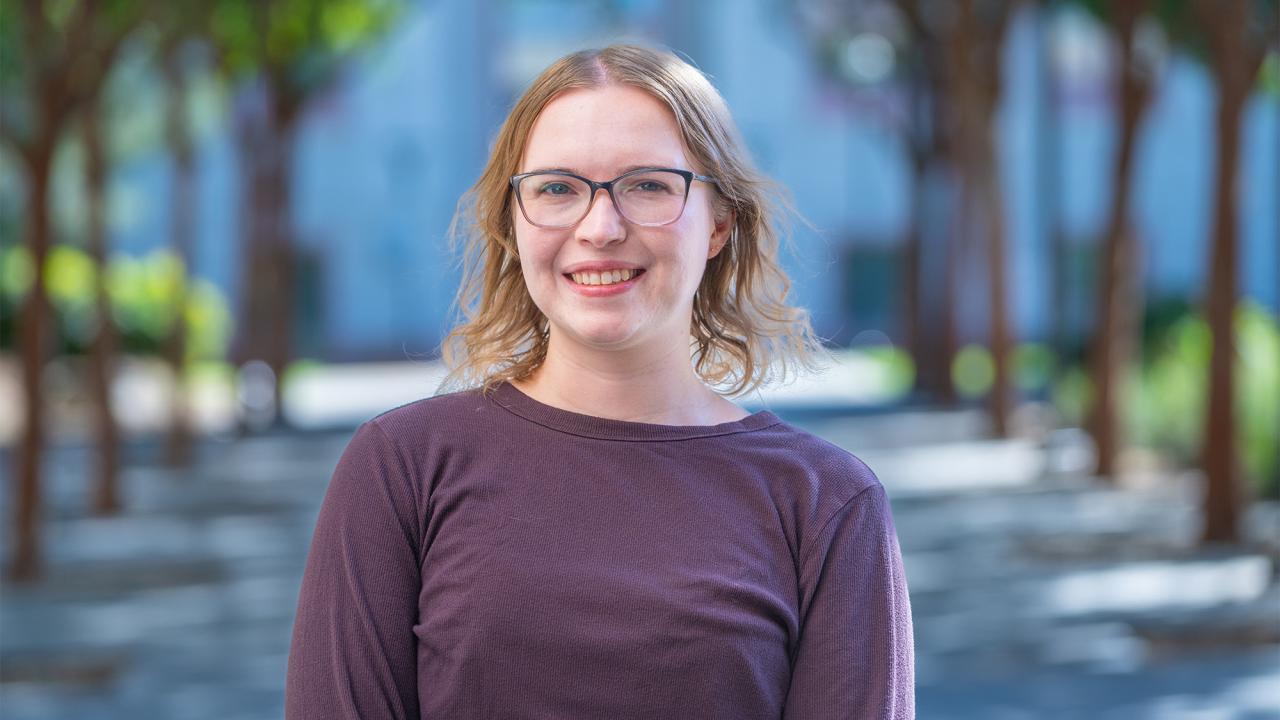 Smiling woman with glasses and short blonde hair in purple top, blurred tree-lined city street.