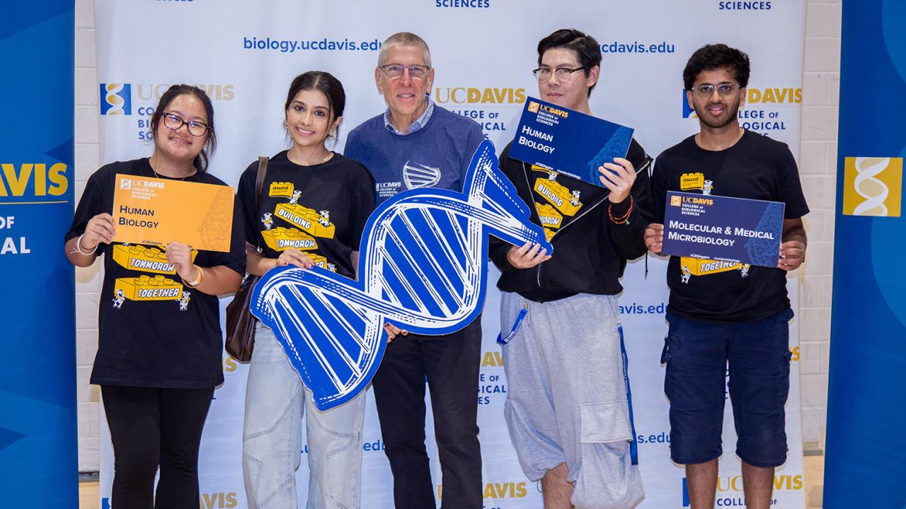 Five people smiling at outreach event, holding large blue DNA prop and signs