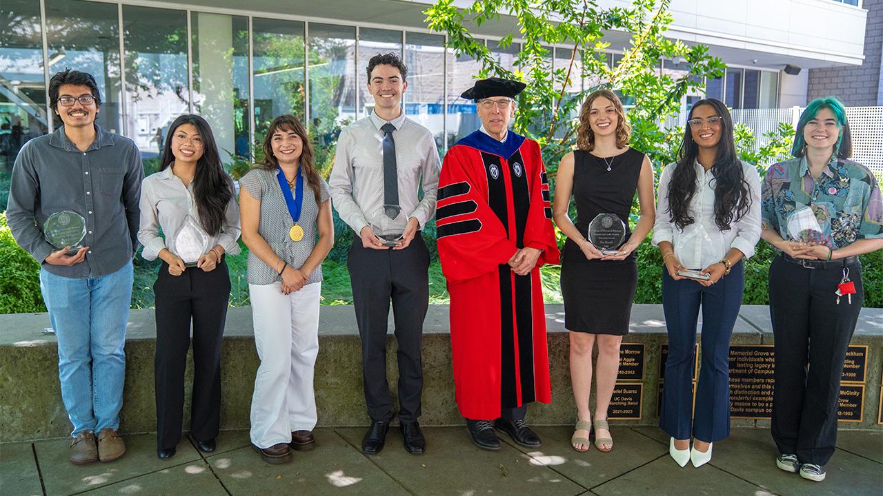Nine people by glass building, center in red academic gown, others in semi-formal attire