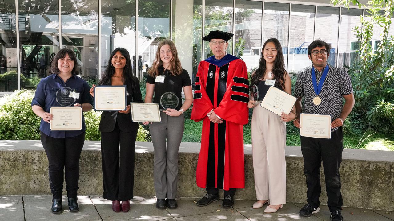 Six people stand in a row outdoors in front of a glass-walled building. Five students, holding awards and certificates, pose on either side of a man in a red and black academic robe and cap. The students appear to be honored at a recognition event.