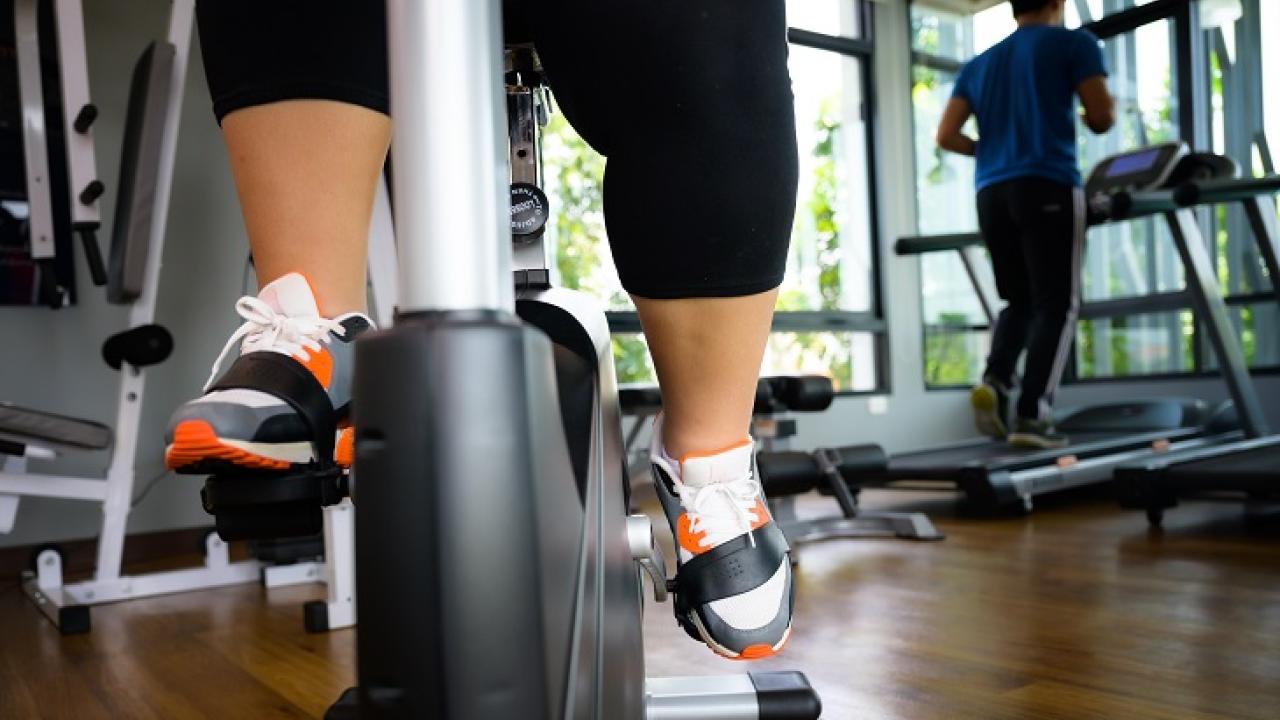 Close-up of a person’s legs pedaling on a stationary exercise bike in a gym, with another individual running on a treadmill in the background.