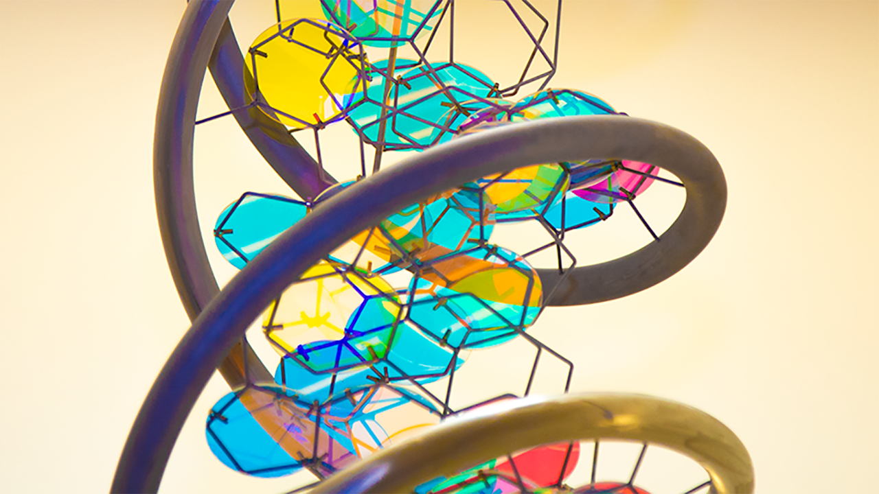 A close-up view of a colorful, abstract sculpture featuring intertwined metallic loops in dark and bronze tones. Within the loops are irregularly shaped panels resembling shards of stained glass in blue, yellow, and pink hues. Thin black lines connect the panels, creating an intricate web-like structure.