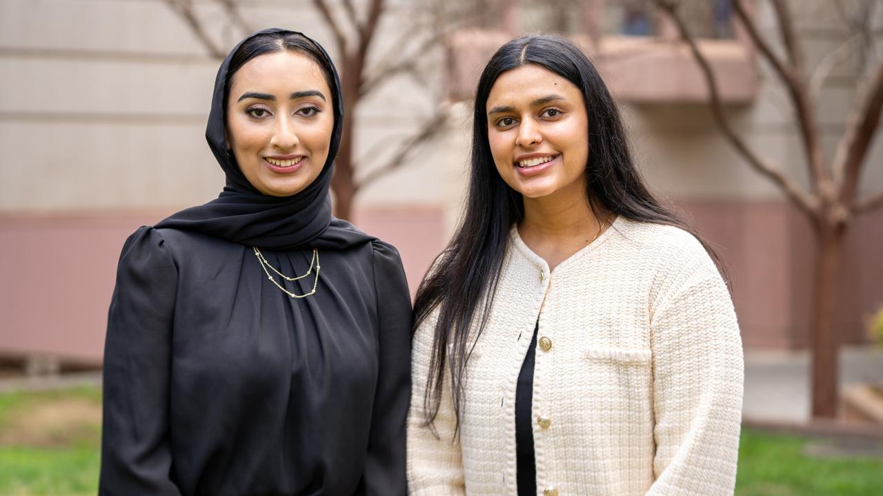 Two young women stand side by side outdoors, smiling at the camera. One wears a black hijab and a black blouse with gold necklaces, while the other has long black hair and wears a cream-colored blazer over a black top. The background features a blurred building and trees.