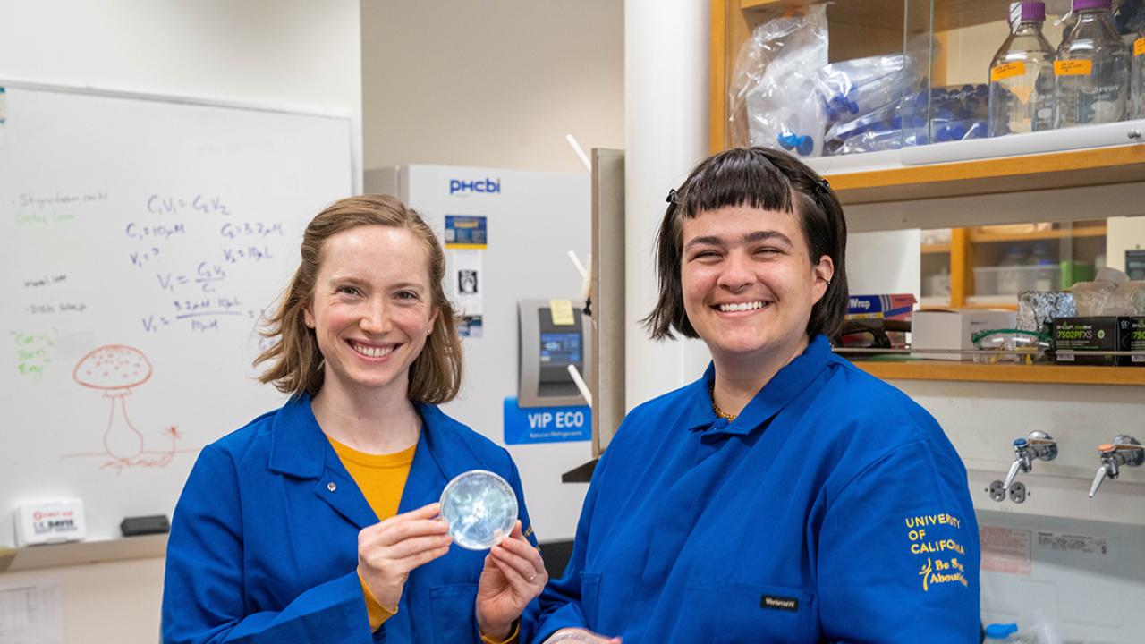 Two researchers in blue lab coats smile in a lab, one holding a petri dish with samples.