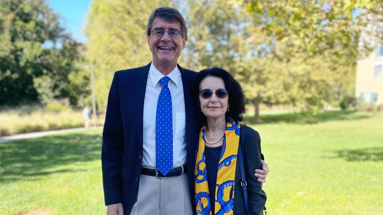 A man and woman stand together outdoors on a sunny day, both smiling slightly. The man wears a dark suit with a white shirt and a blue tie, while the woman wears a dark outfit with a colorful yellow and blue scarf. They appear relaxed, with trees and green grass in the background.