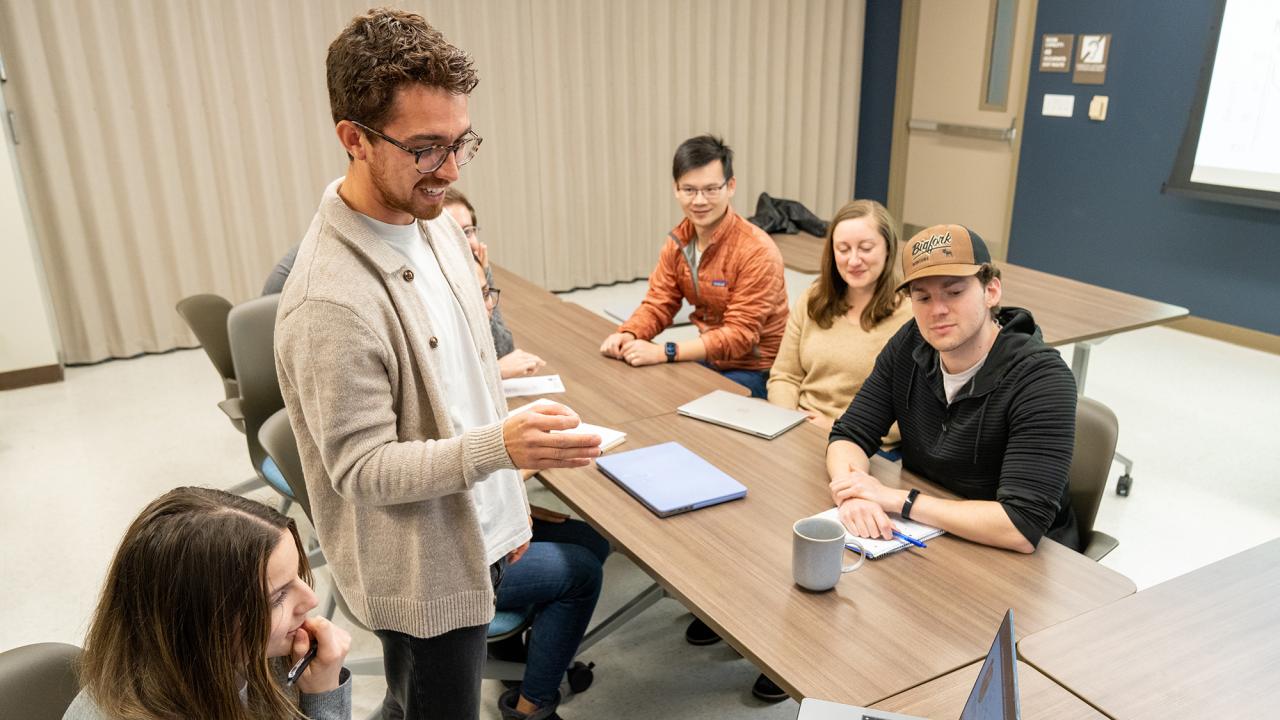 Nate Herrera, a UC Davis graduate student, smiles and gestures while leading a small group discussion in a classroom. Seated students listen attentively, some taking notes, with laptops and notebooks on the table.