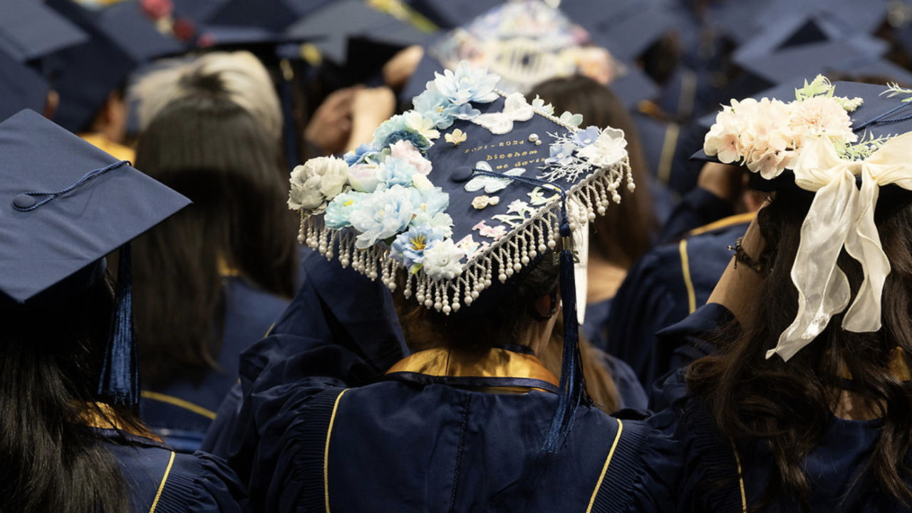 A close-up of a decorated graduation cap adorned with blue flowers, pearls, and a ribbon, surrounded by other graduates in blue and gold academic regalia.