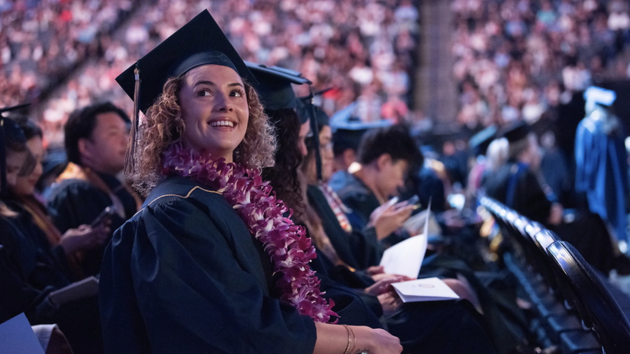 A smiling graduate wearing a flower lei sits among fellow graduates in a large venue, surrounded by a cheering audience.