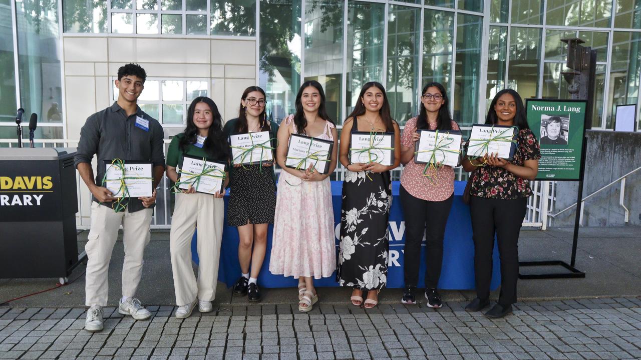 Seven UC Davis students stand in a row holding certificates after receiving the 2025 Norma J. Lang Prize for Undergraduate Information Research. They are smiling and posed in front of the UC Davis Library entrance, with a podium and a poster honoring Norma J. Lang visible in the background.
