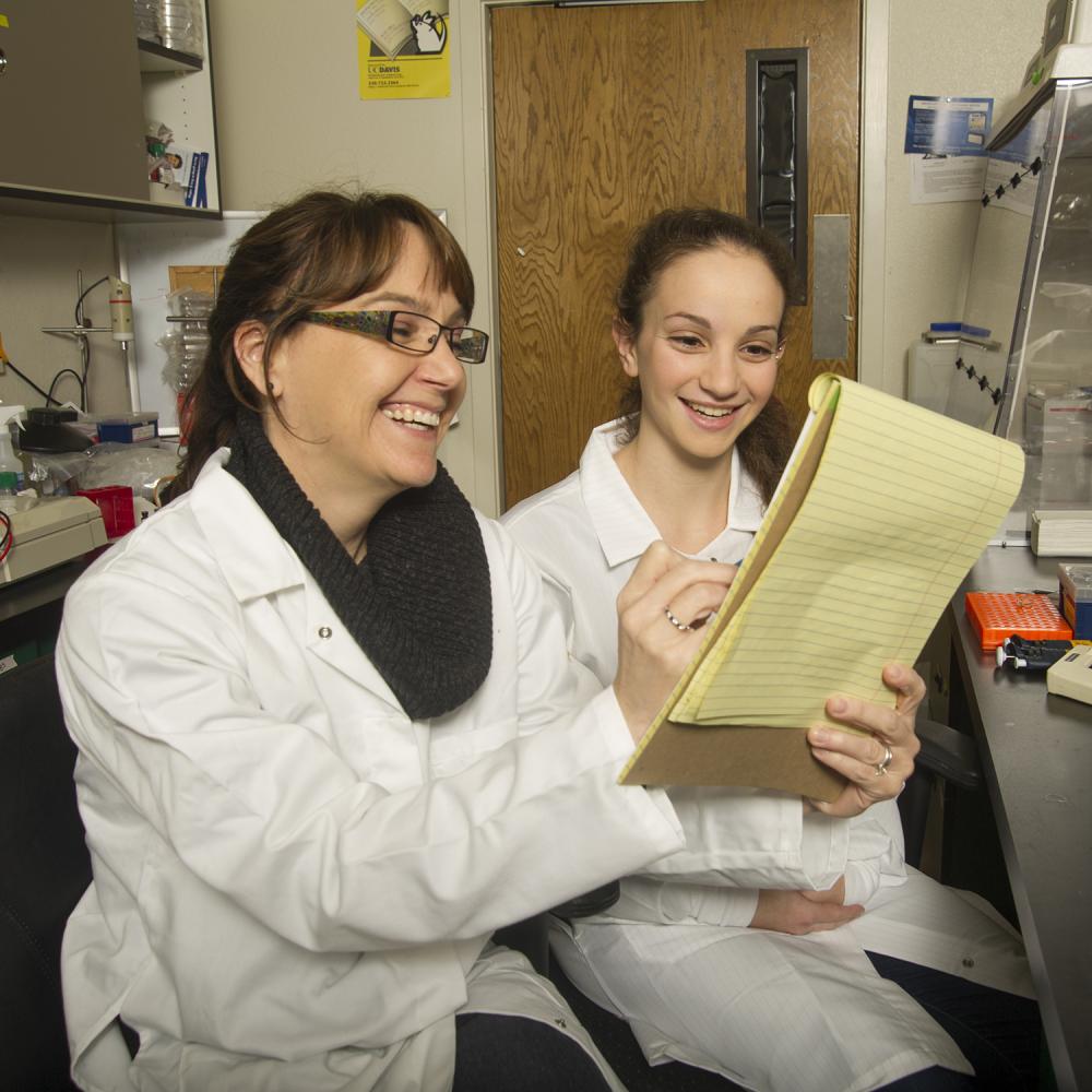 Instructor and student in lab coats smile while reviewing notes together in a biology lab setting.