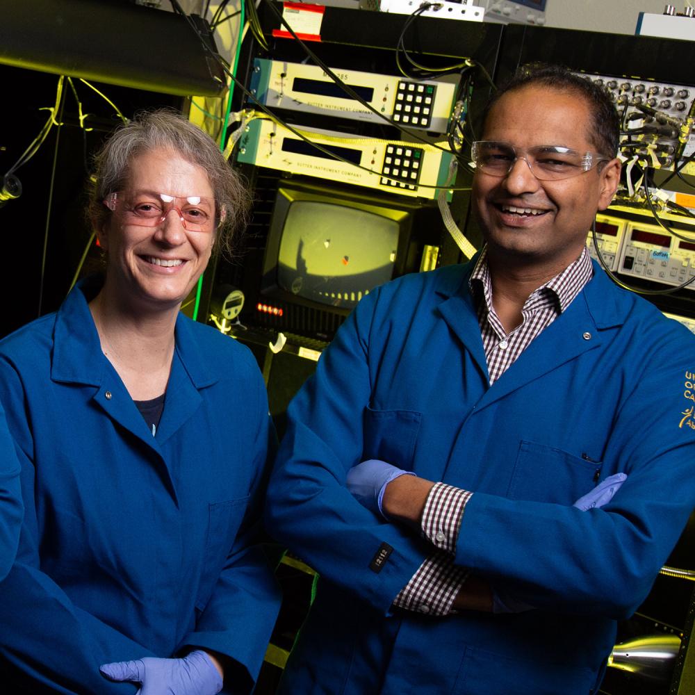 Two researchers in blue lab coats stand smiling in front of complex equipment and monitors in a UC Davis laboratory.