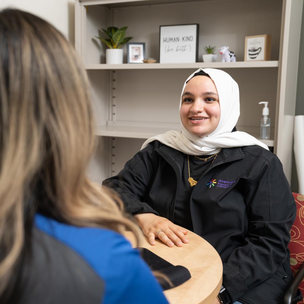Individual person wearing a white hijab smiles at individual with blonde hair facing away from the camera